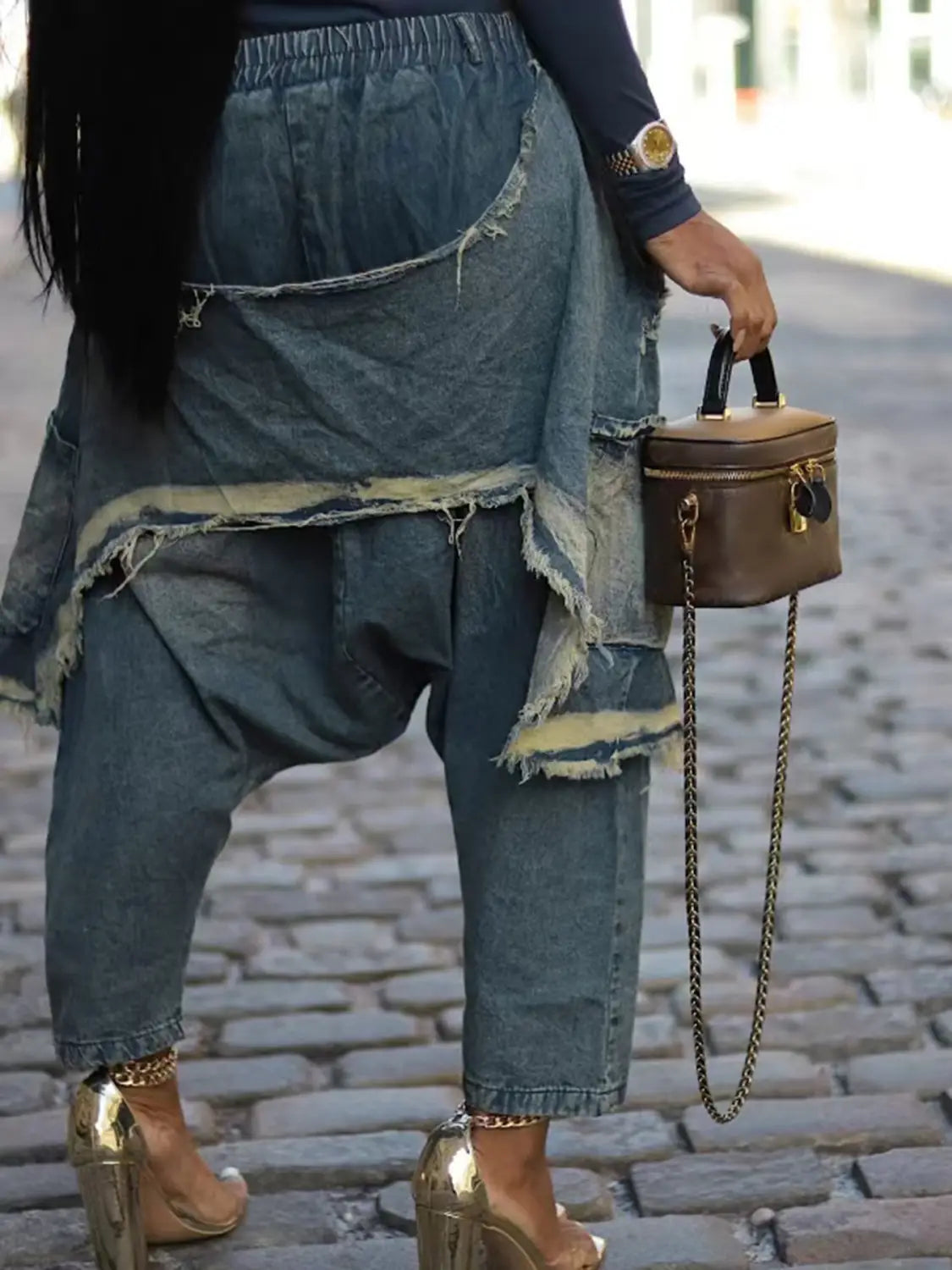Eine kompakte braune Ledertasche aus hellem Tan mit schwarzen Tragegriffen und einem goldfarbenen Kettenriemen, passend zu bestickten Patchwork-Jeans-Haremshosen.