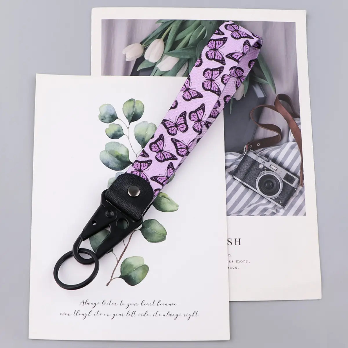 A butterfly wrist strap keychain with a lavender fabric strap featuring a repeating purple butterfly pattern, black plastic clip, and metal ring.