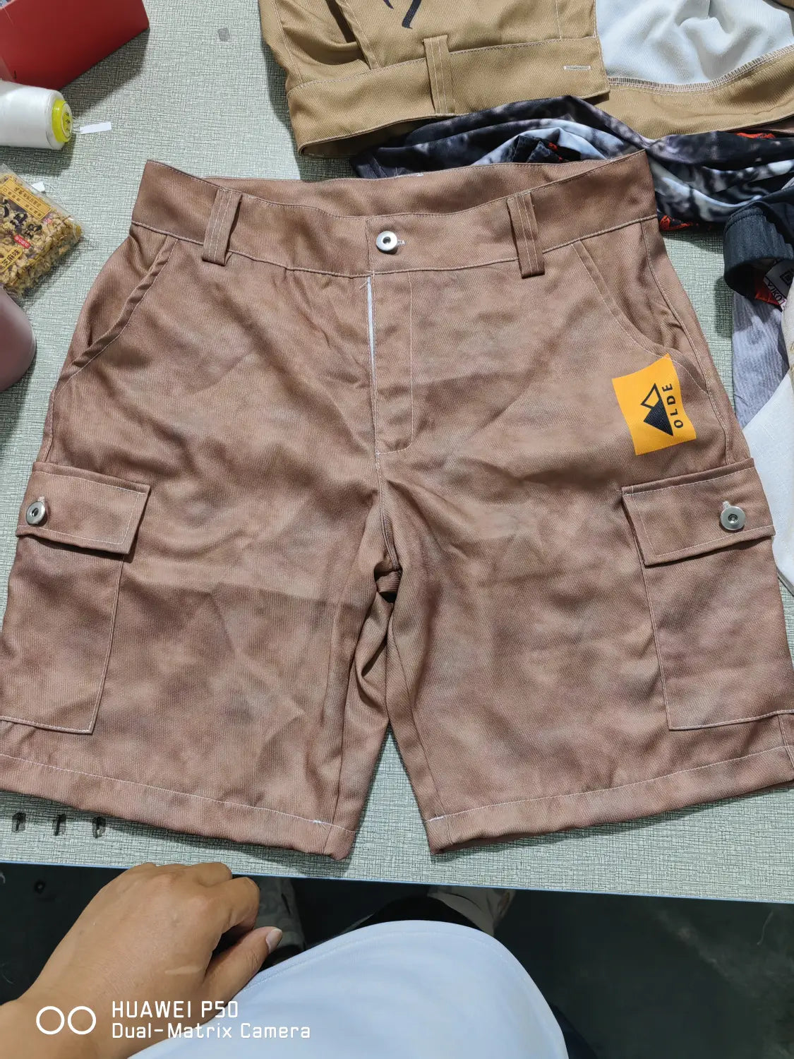 Men’s digital print cargo shorts in tan polyester featuring two large side pockets secured by silver buttons and a yellow brand tag on the right hip.