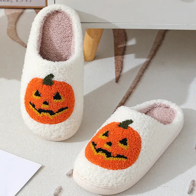White plush jack-o-lantern embroidered Halloween slippers feature a cheerful orange pumpkin face with black triangular eyes and mouth accented by yellow stitching.
