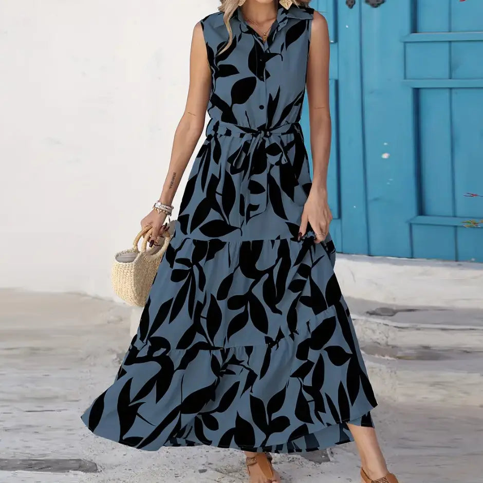 Elegant leaf print sleeveless tiered maxi dress in muted blue fabric with bold black leaf patterns and a self-tie waistband.