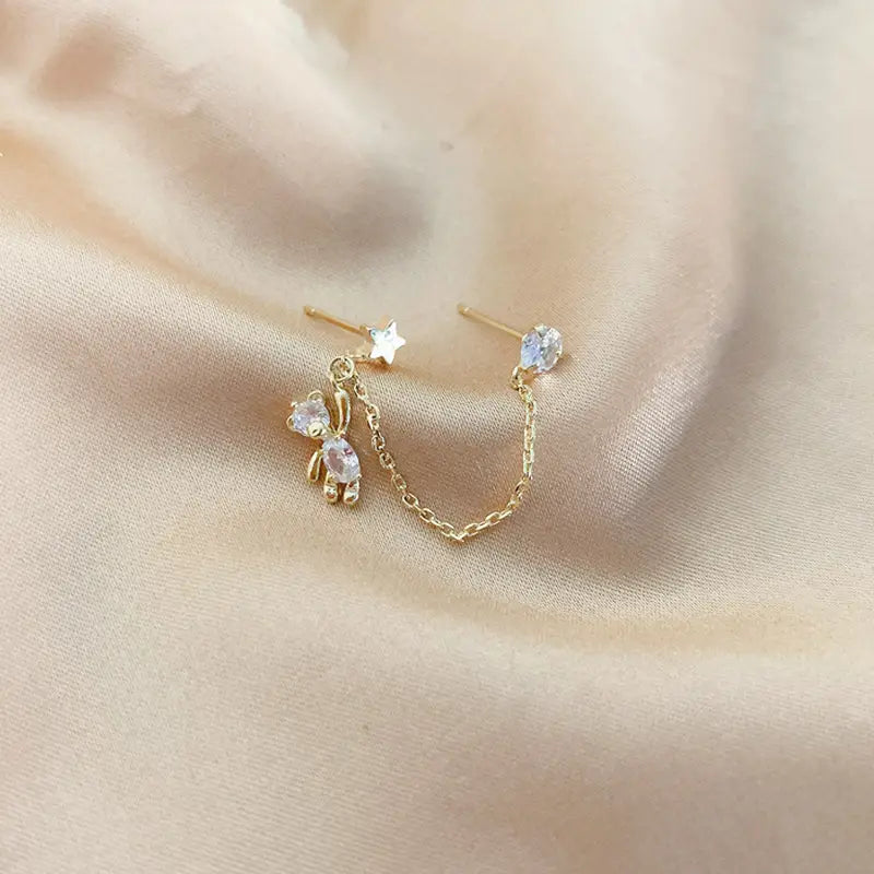Boucles d’oreilles asymétriques en strass à finition dorée, avec ours et étoiles, comportant une pastille en forme d’étoile et une breloque ours suspendue ornée de pierres précieuses transparentes, reliées par une chaîne élégante.