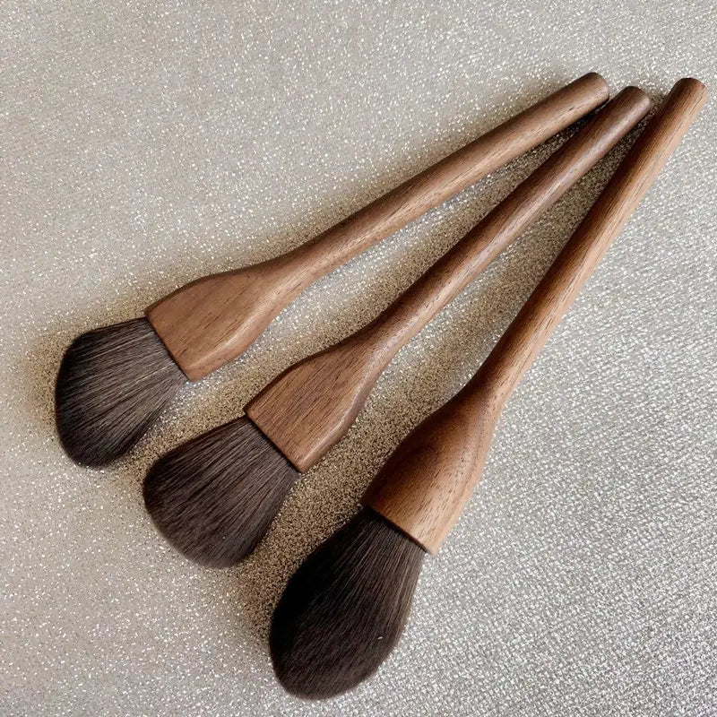 three wooden-handled walnut wood makeup brushes with soft dark brown fiber bristles arranged diagonally on a textured surface