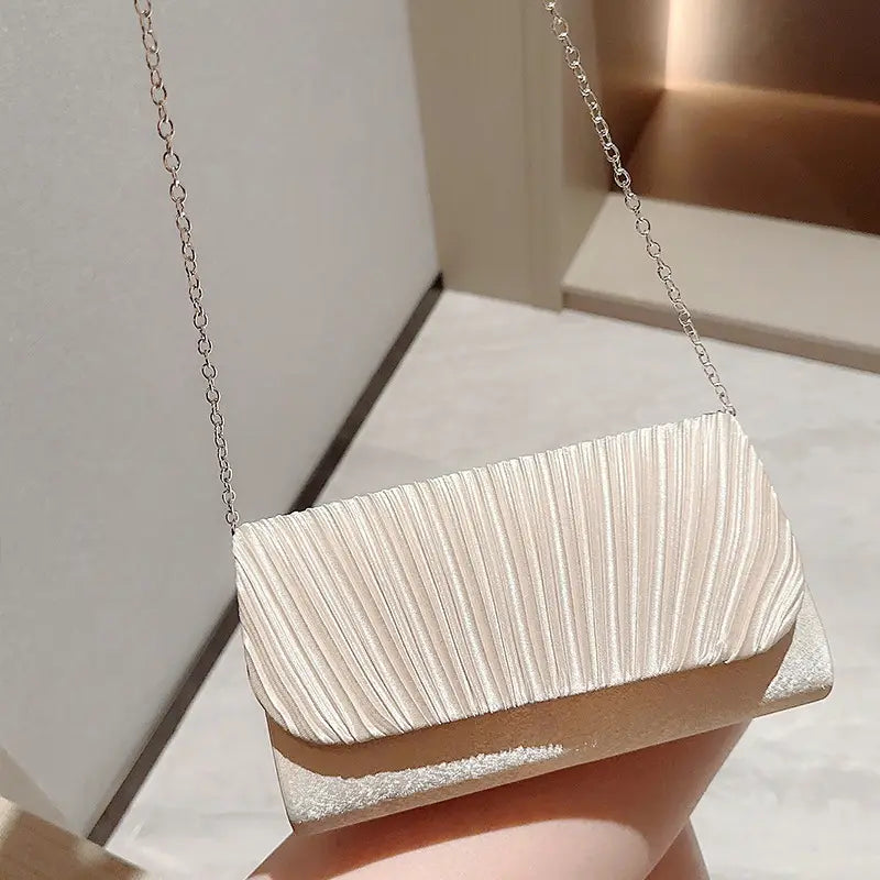 A sleek off-white pleated satin clutch with a metallic silver base and delicate chain strap designed as a minimalist women’s evening bag.