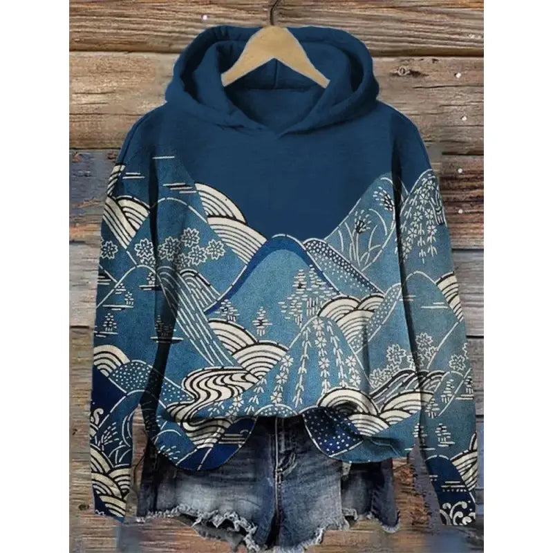 Sudadera con capucha azul con un patrón detallado de olas y montañas en tonos azul claro y blanco, colgada de una percha de madera de la colección de sudaderas con capucha de estampado de olas japonesas.