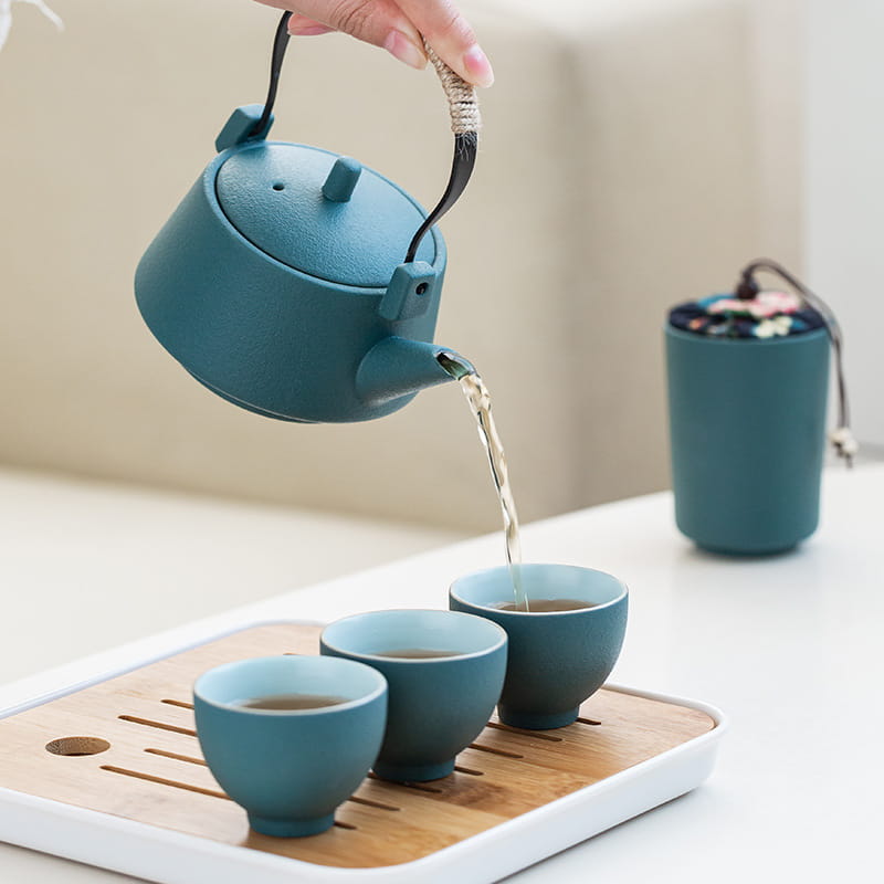 Teal ceramic travel tea set featuring a teapot with a woven rope handle pouring tea into matching teal cups on a bamboo tray with a case.