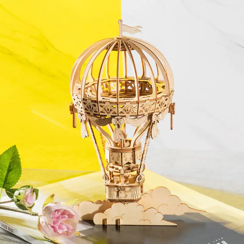 Un kit de construction de modèle mécanique 3D d’assemblage DIY en bois, représentant un ballon à air chaud en bois finement ouvragé, avec des motifs découpés avec précision, un petit drapeau sur le dessus, et un intérieur détaillé incluant une figurine dans le panier.