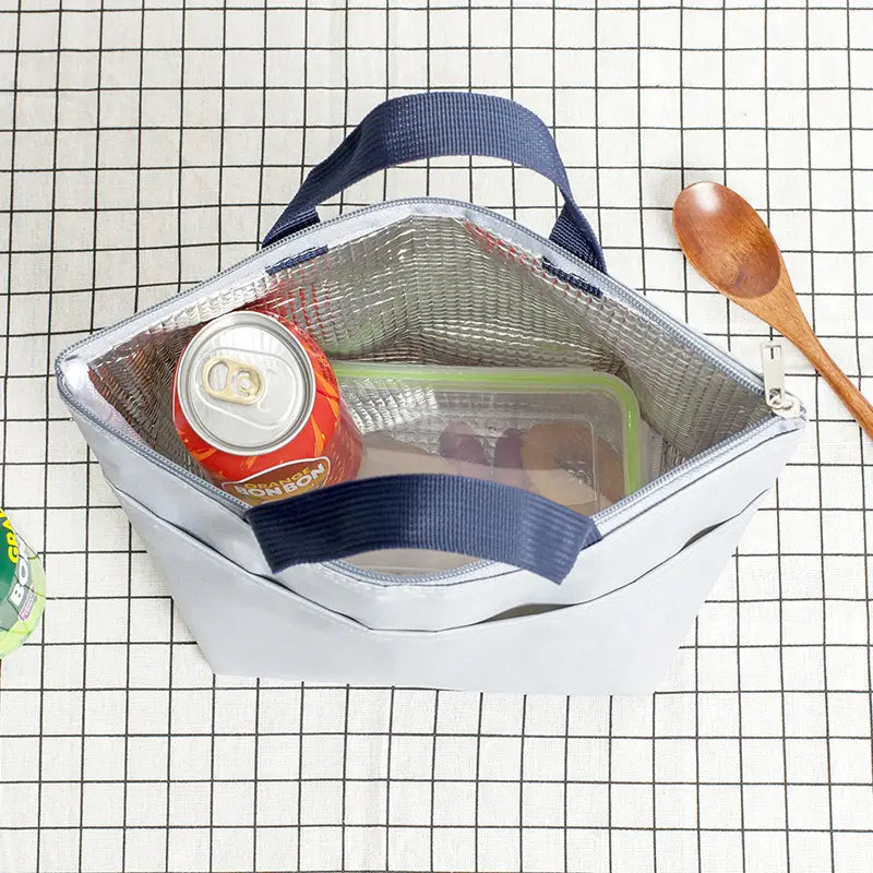 Hellgraue isolierte Oxford-Bento-Lunchbag mit dunkelblauen Stoffgriffen und Reißverschlussverschluss; mit silbernem reflektierendem Innenfutter, das eine Dose Erfrischungsgetränk und einen klaren Behälter enthält.