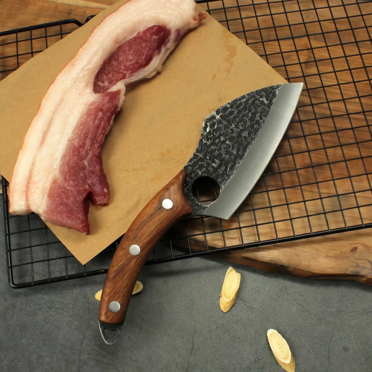 Un couteau à désosser rustique en acier martelé, forgé à la main en acier au carbone à haute teneur, avec un manche en bois brun riche et des rivets en argent, repose à côté d’une tranche de poitrine de porc sur du papier sulfurisé.