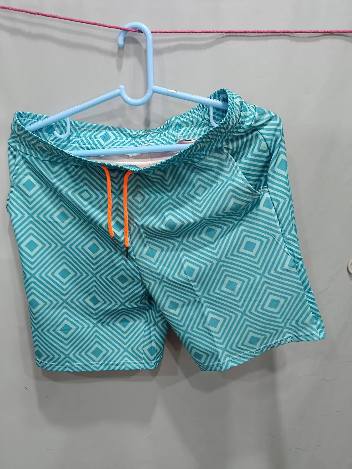 men’s geometric print double layer beach shorts in teal with a geometric diamond pattern and orange drawstrings hanging on a light blue hanger.