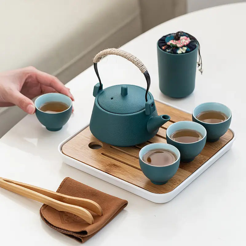 Teal ceramic travel tea set with woven handle bamboo tray and matching cups arranged on a wooden tray.