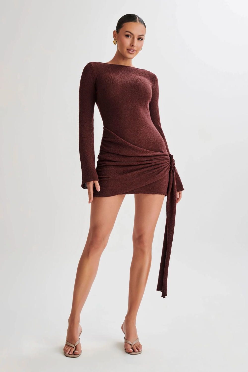 A sleek burgundy long sleeve knit mini dress with a backless design and draped side tie detail featuring a subtle sheen.