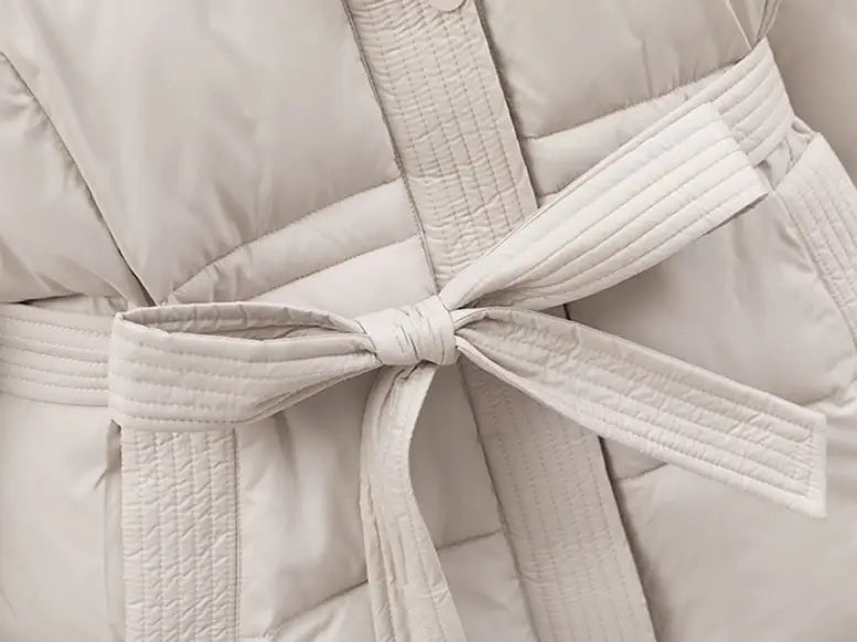 A cream-colored quilted hooded winter puffer coat for women featuring a matching belt tied in a neat bow.