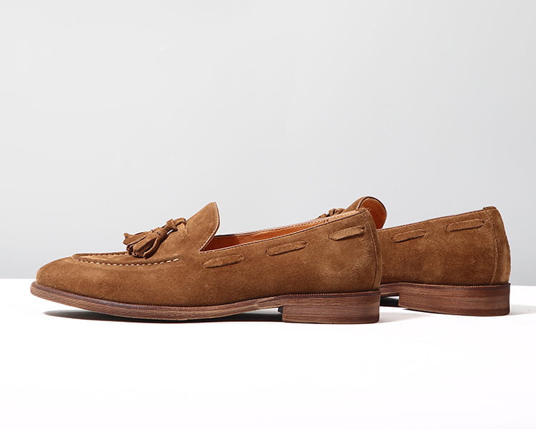 Des mocassins à pampilles en daim beige, avec des coutures cousues et une semelle en caoutchouc cousue, sont présentés dans un gros plan détaillé.