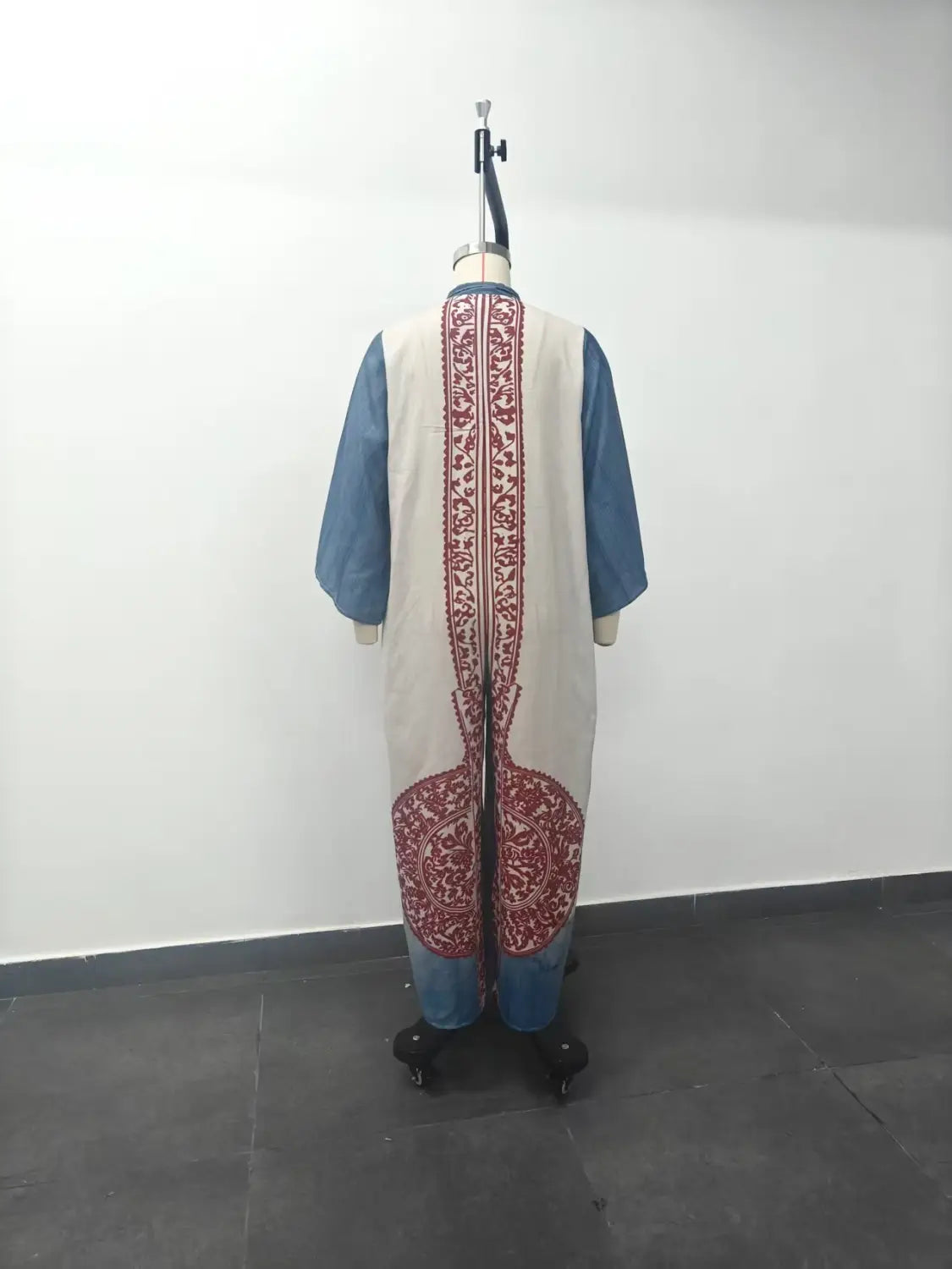 Un gilet long de couleur crème, brodé de motifs floraux rouges complexes au dos et à l’ourlet, porté par-dessus une chemise bleue, associé à un pantalon bohémien à jambes larges imprimé.