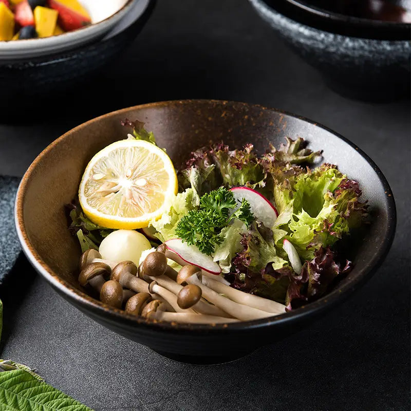 A rustic dark brown ceramic ramen bowl filled with a fresh salad of mixed greens radishes mushrooms and lemon slice, ideal speckled black blue kitchenware.