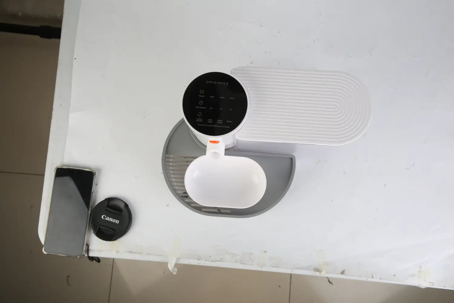 Fontaine à eau intelligente pour animal de compagnie blanche et grise sans fil rechargeable, avec un corps en plastique élégant, un panneau de commande circulaire noir et une gamelle ovale pour l’alimentation automatisée de l’animal.