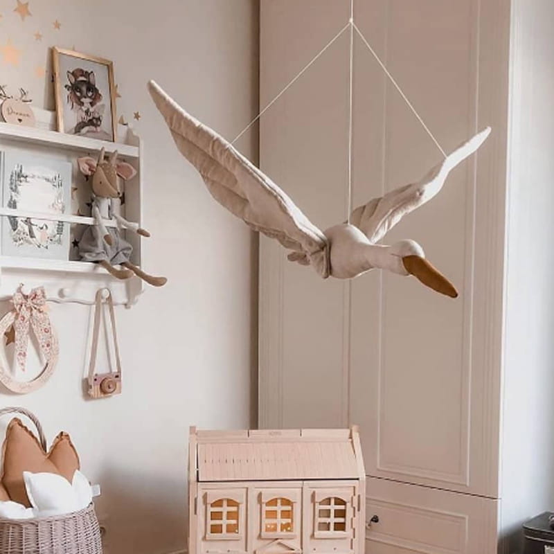 Mobile en peluche souple, blanc cassé, représentant une cigogne, avec un bec beige, suspendu par des cordelettes dans une chambre d’enfant, pour servir d’oreiller d’animal en forme de cygne blanc.