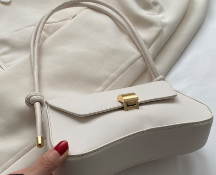 Minimalist faux leather structured shoulder bag in cream color featuring a gold-tone clasp and matching drawstring handle.