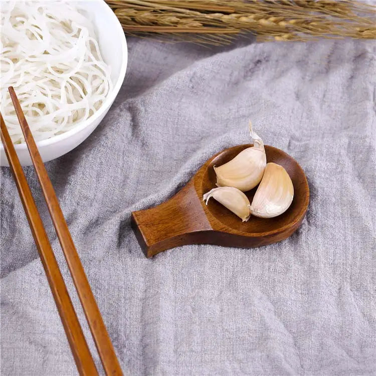 Plato de madera para salsa de inmersión con soporte para palillos, que presenta una pequeña cuchara de madera pulida con un cuenco redondeado y liso, que sostiene dientes de ajo pelados.