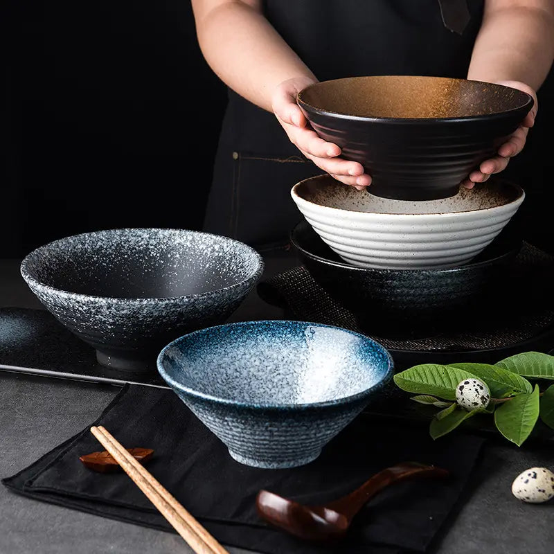 A set of ceramic ramen bowls with speckled glazes and ribbed exteriors featuring a black bowl held above stacked white and dark blue bowls for kitchenware use.