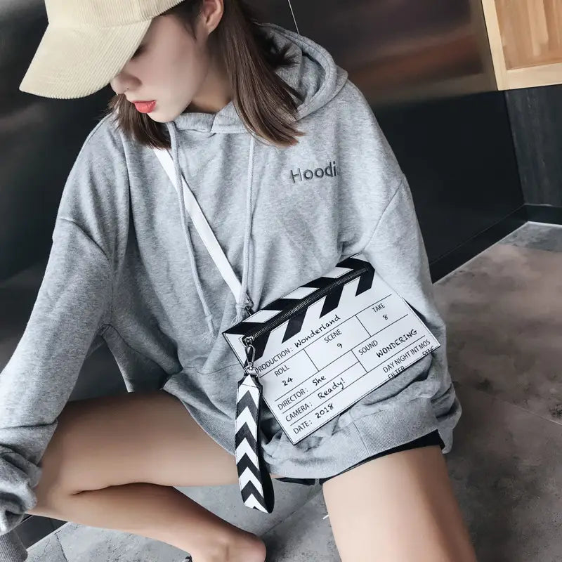 Une personne portant un sweat à capuche gris, avec un logo brodé sur la poitrine et un cordon de serrage blanc, tient une pochette enveloppe bandoulière en cuir PU représentant un clap de cinéma.