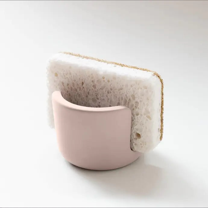 A soft pink ceramic diatom mud sponge rack holding a white textured cleaning sponge with a golden-brown abrasive edge for use as a sink caddy holder.