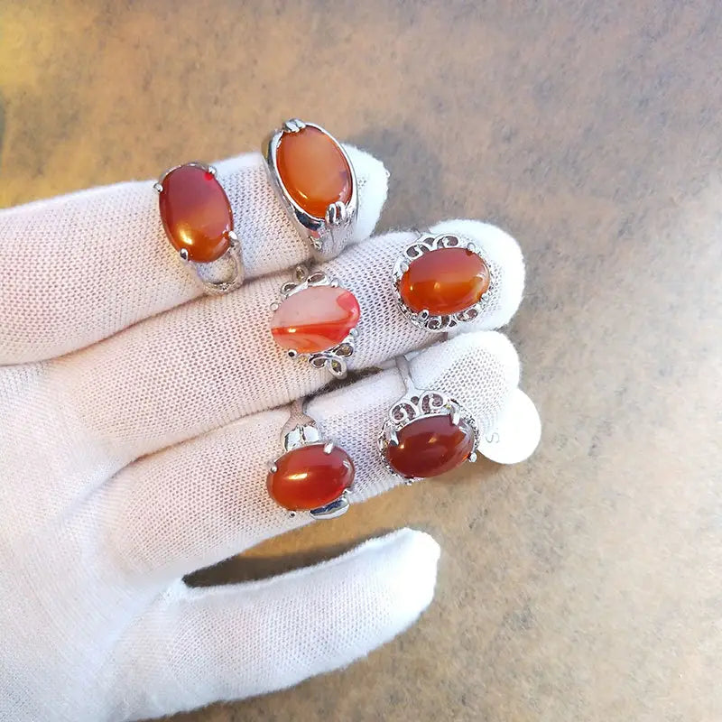 A hand in a white glove displays five oval-shaped carnelian natural gemstone silver tone statement rings with metal settings.