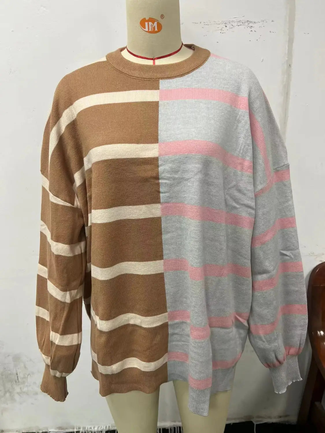 a women’s oversized knit sweater with a split-color striped design featuring warm brown and cream stripes on one side and soft gray and pink stripes on the other, long sleeves, and ribbed cuffs
