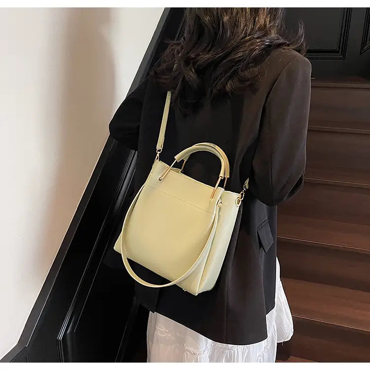 Cream-colored minimalist leather bucket bag with gold-tone hardware, a top handle, and a detachable strap.
