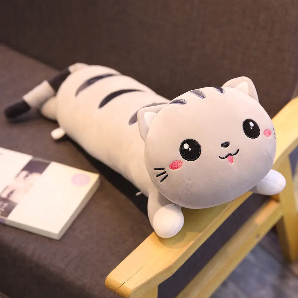 A soft plush white cat with black stripes and pink cheeks lies stretched out on a wooden surface as a stuffed animal toy pillow.