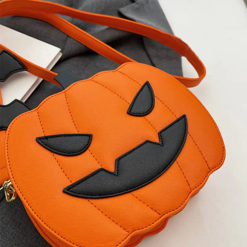 Orange pumpkin-shaped crossbody bag with black stitched eyes and mouth, quilted texture, and matching orange strap, featuring a detachable bat charm.