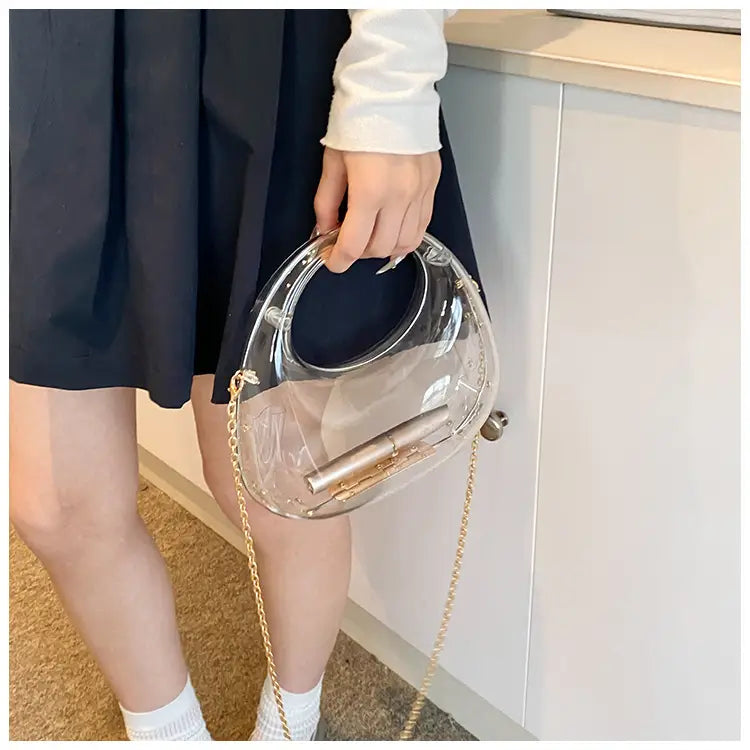 Transparent circular acrylic handbag with a gold chain strap and metallic handle designed as a clutch bag.