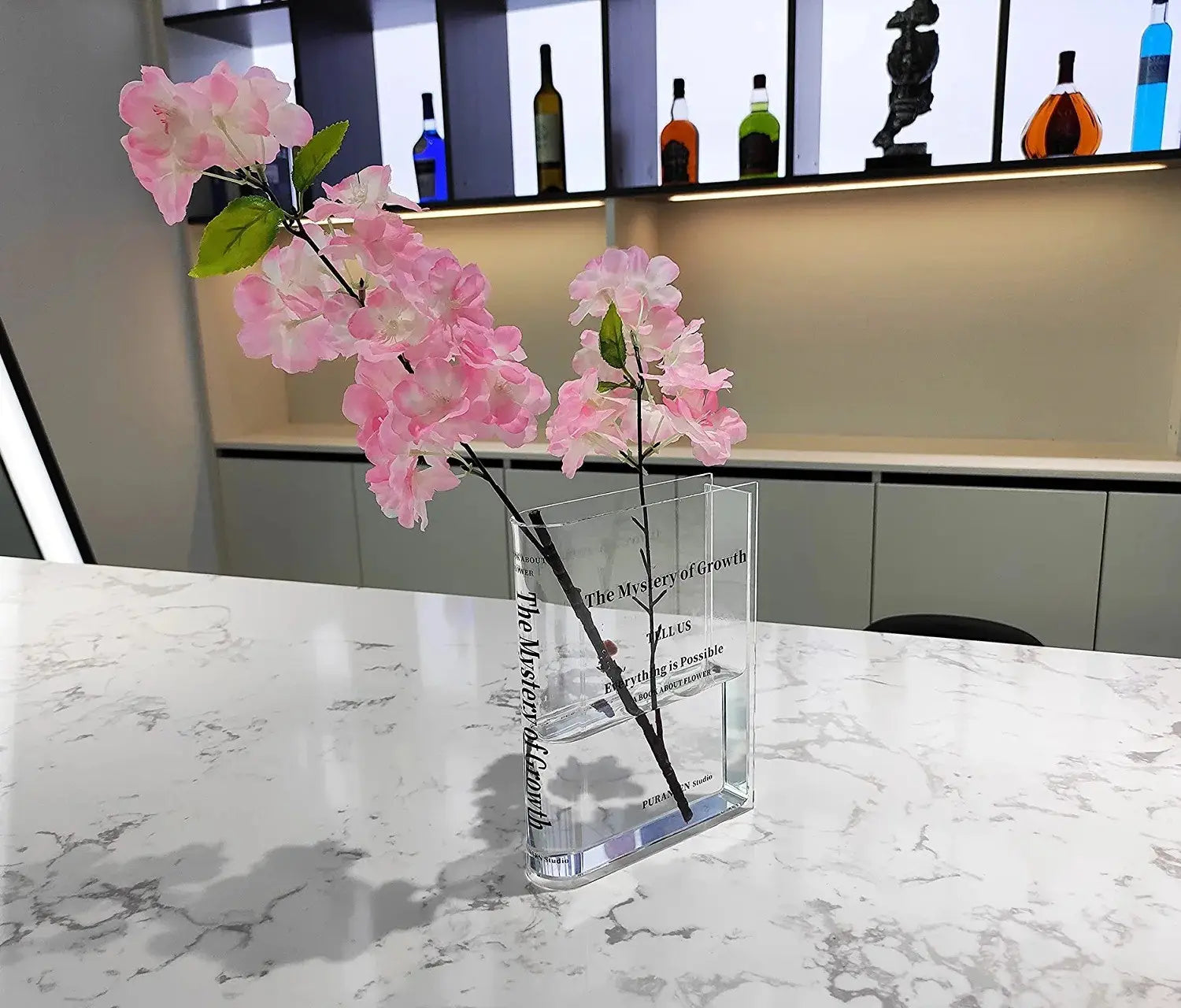 jarrón de flores de libro de acrílico transparente con un diseño rectangular moderno y texto en relieve en los laterales, que sostiene delicadas flores artificiales de color rosa para decoración del hogar.