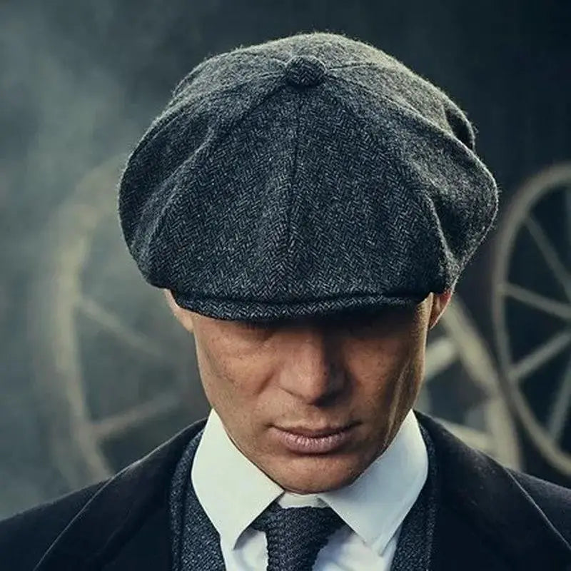 A dark gray herringbone tweed flat cap with an octagonal beret design featuring a rounded crown and stitched button on top.