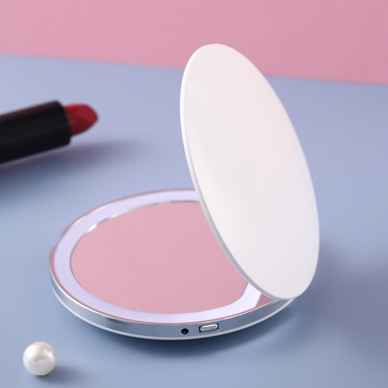 Un espejo de maquillaje LED metálico plateado y redondo, elegante, con tapa blanca e interior iluminado en color rosa, diseñado como espejo portátil USB compacto plegable.