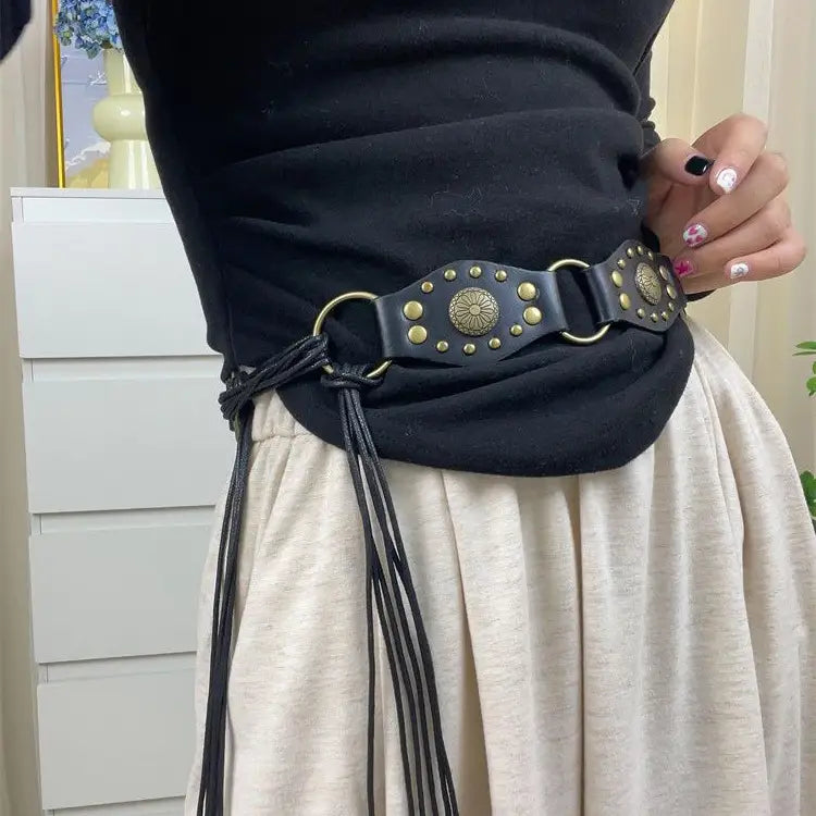Schwarzer Gothic-Punk-Lederharness-Gurt mit goldfarbenen Metallschienen, zentralem kreisförmigem Medaillon und geflochtenen Quasten für einen edgy-böhmischen Look.