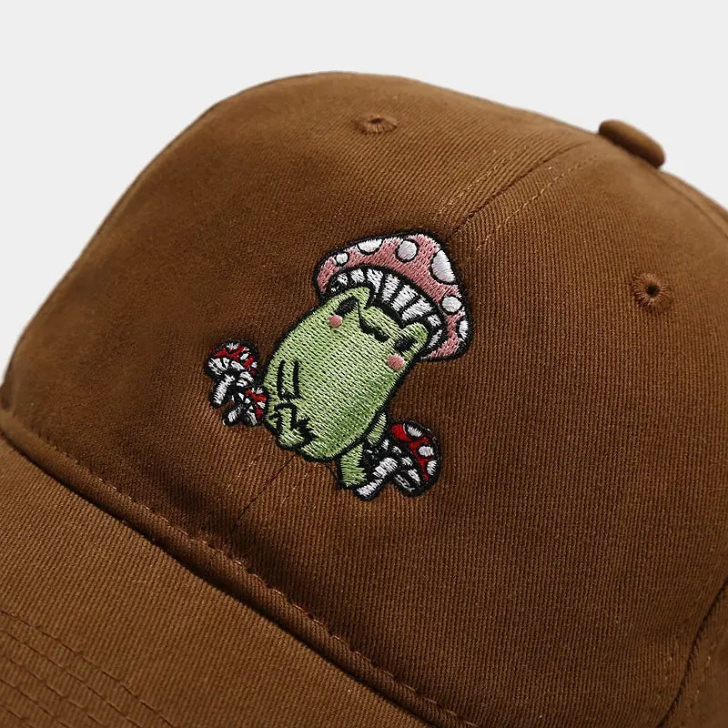 Brown baseball cap made of fabric featuring an embroidered green frog wearing a pink and white mushroom hat surrounded by red-spotted mushrooms titled Frog Mushroom Embroidered Baseball Cap.