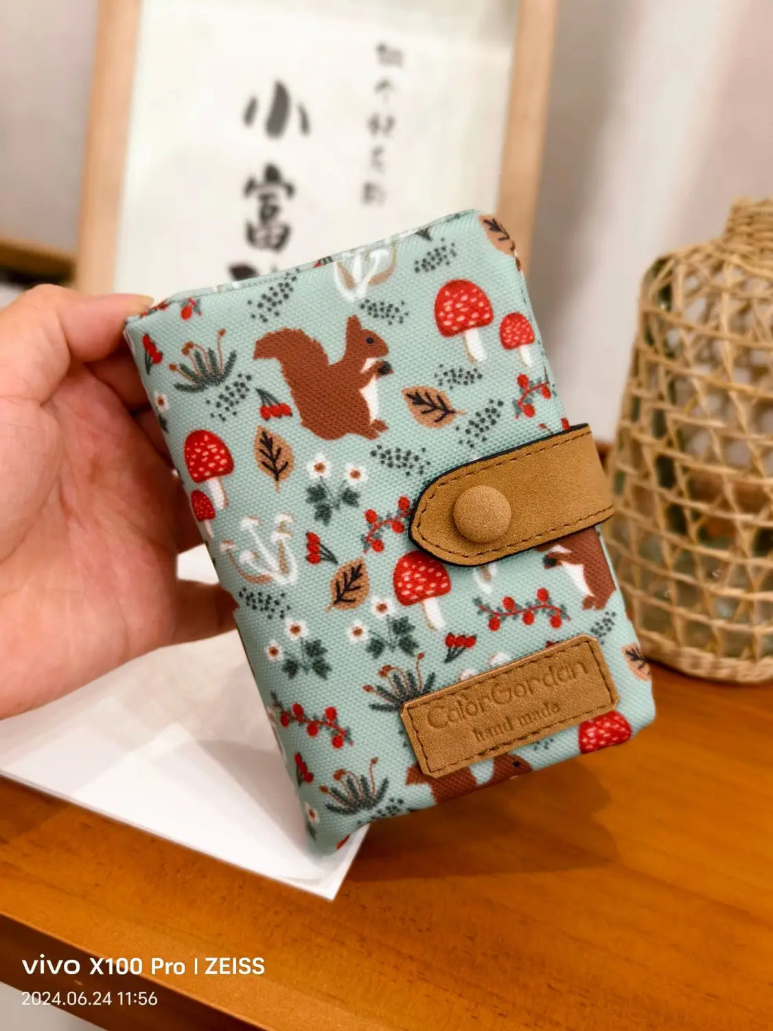 A light blue canvas tri fold multi card wallet with a whimsical pattern of red mushrooms brown squirrels and green foliage accented with tan leather straps and a matching brand patch.
