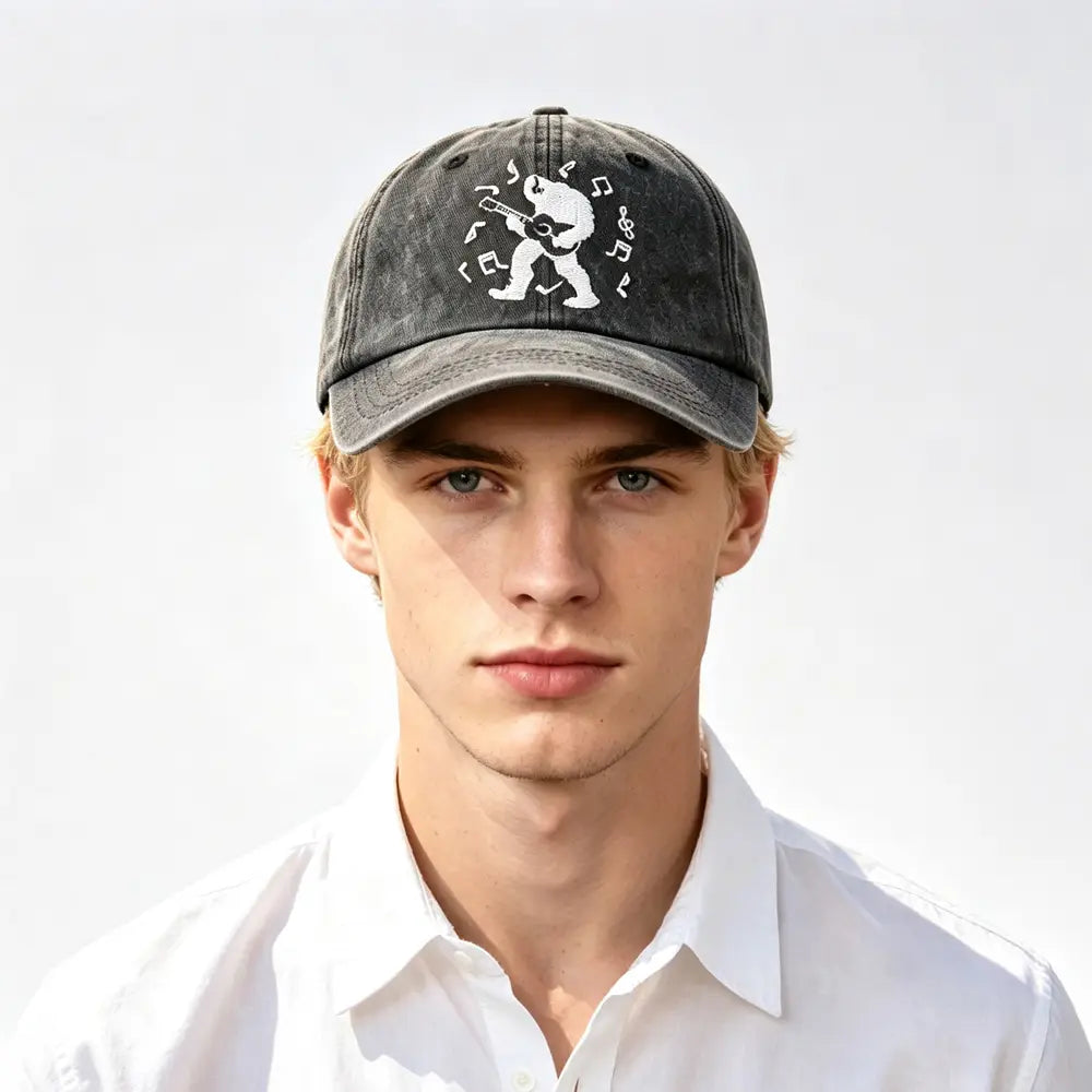 A dark gray distressed denim baseball cap featuring a white embroidered design of a bear playing guitar surrounded by musical notes in the Bigfoot Guitar baseball cap vintage distressed hat.