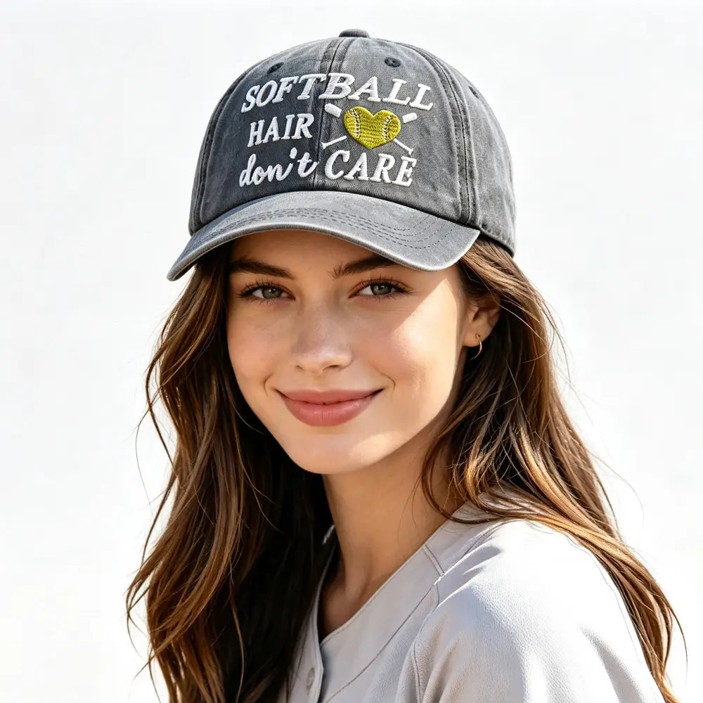 A gray distressed baseball cap featuring white embroidered text reading softball hair don’t care and a yellow heart-shaped softball design.
