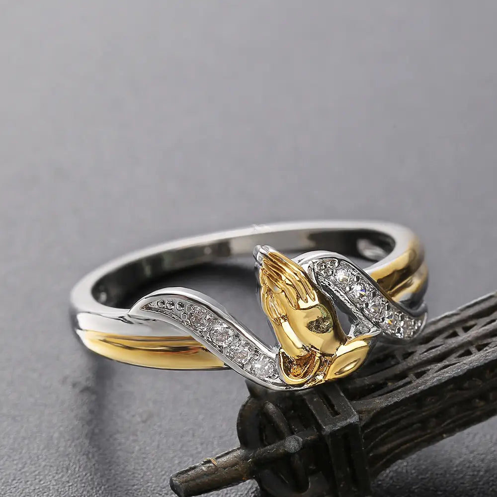 Two tone 18k gold and silver ring featuring a central praying hands design adorned with clear cubic zirconia stones.