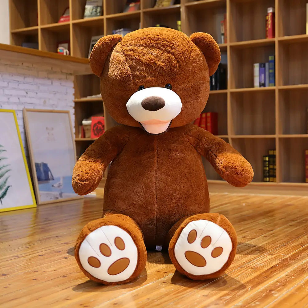 Ein großer Plüsch-Teddybär aus weichem Samt in Braun mit weißen Pfotenpolstern und einer angenähten Nase, der aufrecht auf einem Holzboden sitzt.