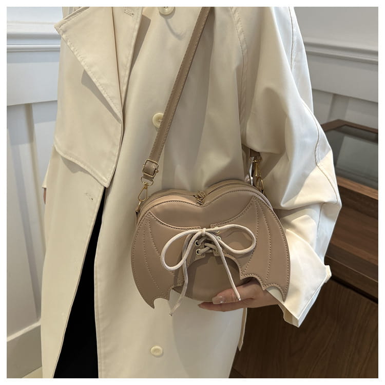 Beige bat wing heart crossbody bag with lace up detail and adjustable strap made of smooth material.