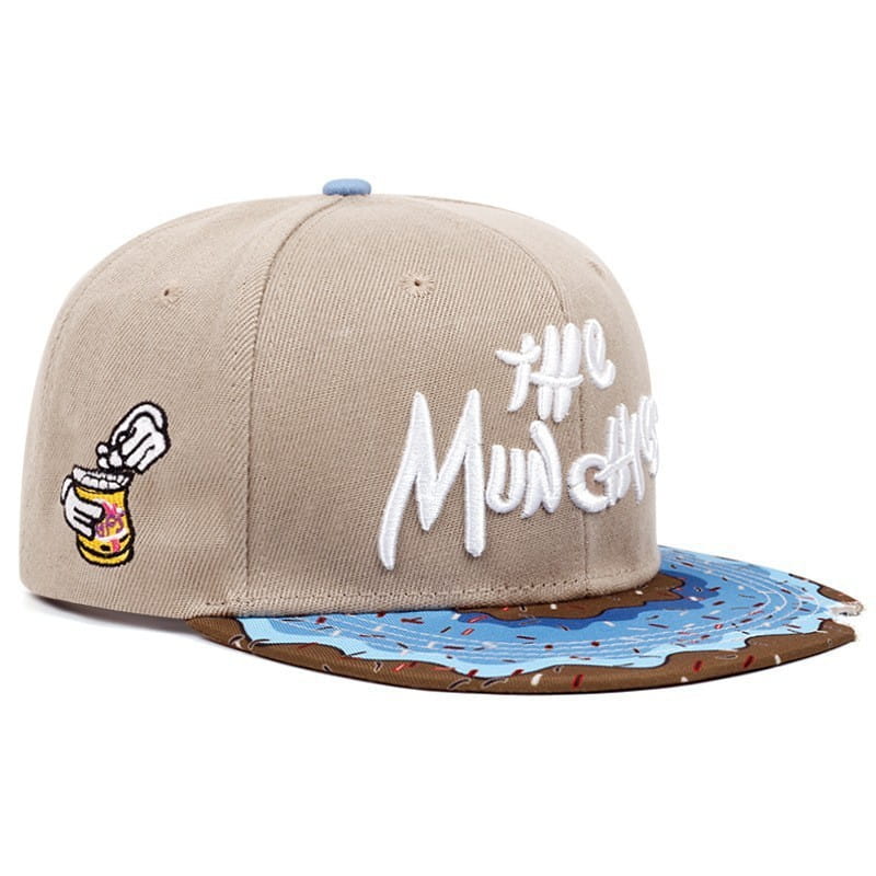 Beige snapback cap with white embroidered the munchies text, a cartoon hand holding a jar patch on the side, and a blue frosting and chocolate sprinkle flat brim.