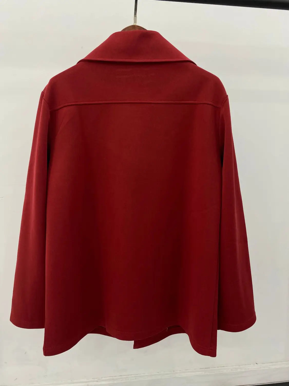 Un abrigo tipo gabardina cruzado doble de algodón rojo granate intenso con cuello estructurado y mangas largas, colgado de forma ordenada en una percha de madera.