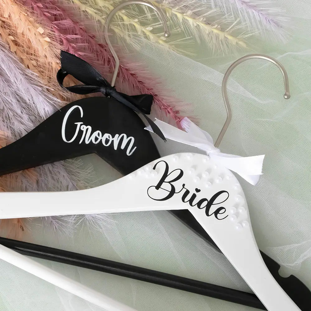 Ensemble de cintres de mariage perles en bois noir et blanc pour la mariée et le marié, avec un cintre portant la mention « marié » et orné d’un nœud en ruban noir, et l’autre portant la mention « mariée », agrémenté d’ornements en perles et d’un nœud en ruban blanc.