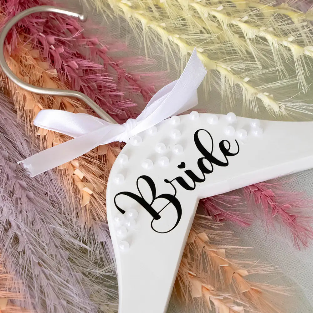 Cintre de mariage de la mariée en bois blanc, orné de perles et d’une inscription cursive noire, noué avec un ruban en satin blanc, provenant de l’ensemble de cintres de mariage perles pour la mariée et le marié.
