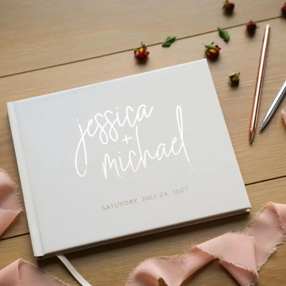 滑らかな表紙にミニマルなデザインで、「Jessica Michael」と「Saturday July 24 2027」と読める上品なゴールド箔レタリングを施した、エレガントな筆記体の文字を特徴とする、パーソナライズ硬表紙の白いカスタム結婚式ゲストブック。