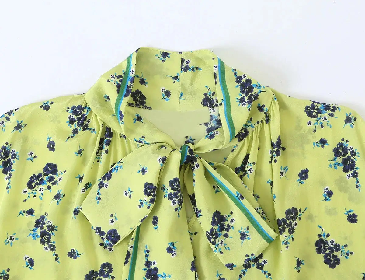 Eine hellgrüne florale Bluse mit zarten dunkelblauen und türkisfarbenen Mustern mit einem weichen Bindekragen-Detail und passender, gestreifter Zierkante aus dem floralen zweiteiligen Midi-Rock-Set im Boho-Outfit-Stil.